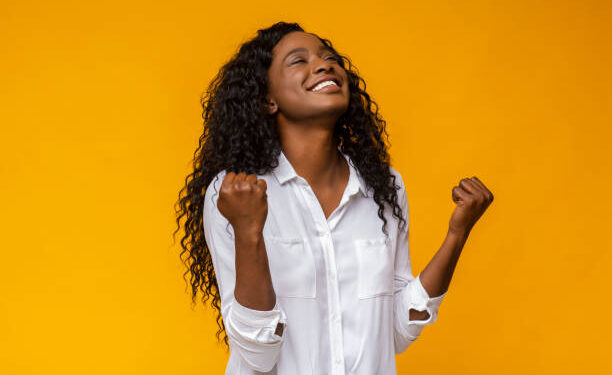 Emotional african american woman smiling and raising clenched fists in the air, feeling excited, yellow studio background