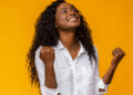 Emotional african american woman smiling and raising clenched fists in the air, feeling excited, yellow studio background