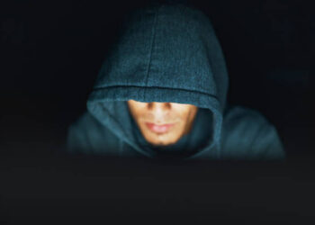 A young man, face partly obscured by a hooded shirt, works on an unseen but illuminated computer by night.