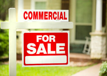 Red and white "Commercial for Sale" sign in front of a stone, wood building that is a residential property. Green grass and bushes indicate the spring or summer season. Front porch and windows in background. Real estate sign in commercial, business district. Relocation concepts.