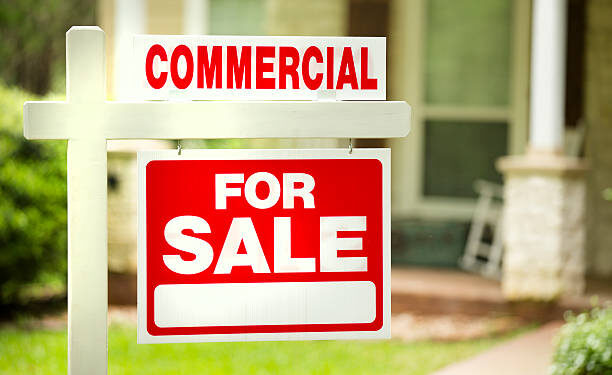 Red and white "Commercial for Sale" sign in front of a stone, wood building that is a residential property. Green grass and bushes indicate the spring or summer season. Front porch and windows in background. Real estate sign in commercial, business district. Relocation concepts.