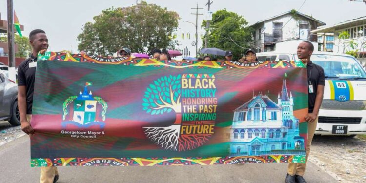 Two students carry the Georgetown Mayor and City Council banner during the Black History Month March on February 27, 2026, proudly displaying the message “Black History: Honouring the Past, Inspiring the Future” as participants proceeded from City Hall to Independence Park.